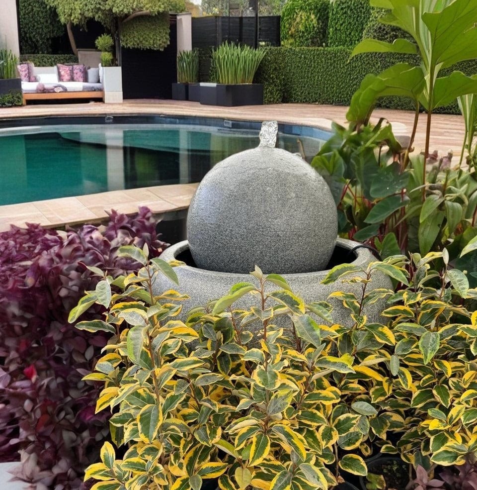 A grey concrete sphere water feature surrounded by colourful foliage beside a pool